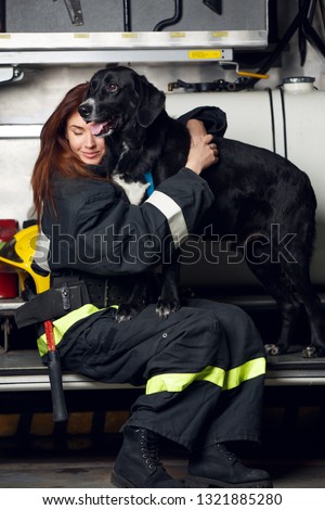 Picture of young woman firefighter with black dog sitting on background of fire truck