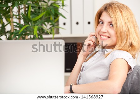 smiling cute young office worker sitting and talking on cell phone in office