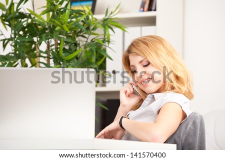 smiling woman sitting, speaking on phone and looking at her watch in the office