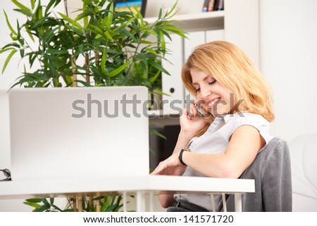 smiling woman sitting, speaking on phone and looking at her watch in the office