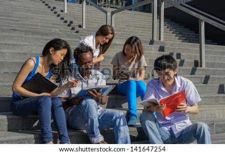 Multiethnic group of university students sitting on steps of university reading notes, metaphor for diversity and international friendship