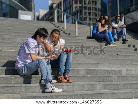 Asian And European Students, Handsome Boy And Beautiful Girl, Sitting On Steps Outside University With Notes And Reading With Several Other Students On Background