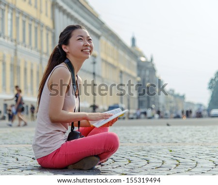 Tourism And Travel Concept. Young Tourist, Beautiful Asian Girl Sitting On Pavement Studying A Map In The Center Of St.-Petersburg. Image Is Professionally Retouched.