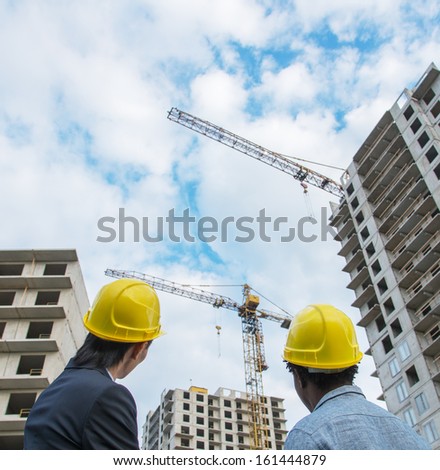 Back View Of Asian And African Designer Or Architect And Foreman Wearing Yellow Hardhats And Looking On Construction Site.