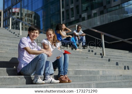 Asian And European Students Sitting On Steps Outside University With Notes And Reading With Several Other Students On Background