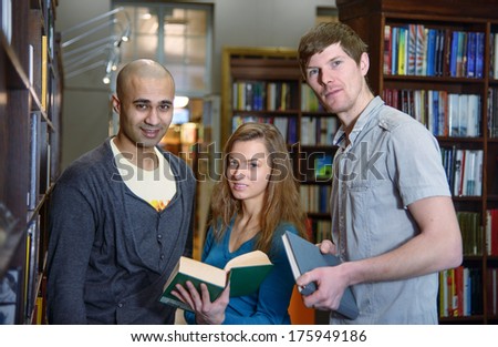 Portrait of three students, european girl and middle eastern and european boy,  standing in a library by a bookshelf and looking at camera