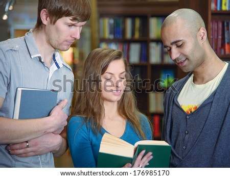 Portrait of three students, european girl and middle eastern and european boy,  standing in a library by a bookshelf and discussing a book