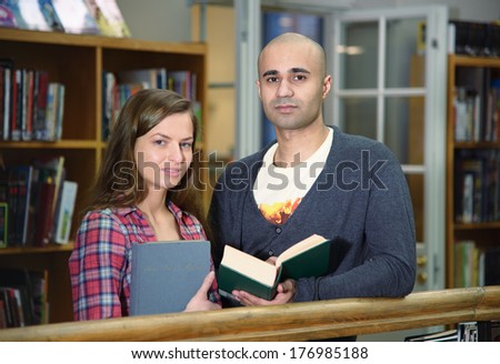 Portrait of two students, european girl and middle eastern boy, standing with books in university library and looking at camera