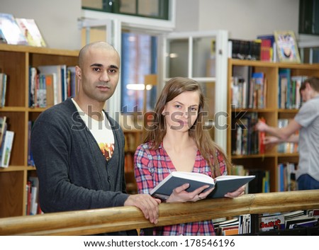 Portrait of two students, european girl and middle eastern boy, standing with books in university library and looking at camera