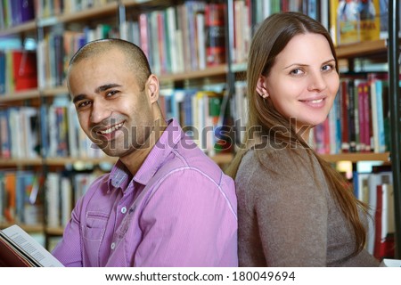 Two young university students sitting back to back in a library and smiling at camera