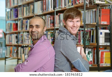 Two young university students sitting back to back in a library and smiling at camera