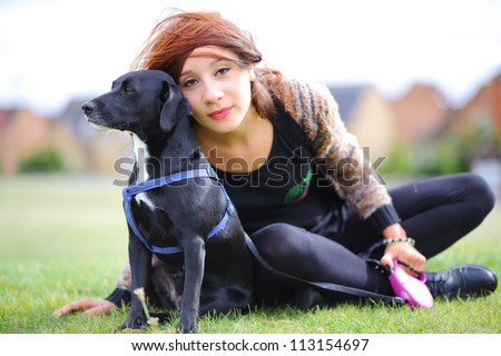 Pretty girl with small black dog looking to side. Suburban scene, in park with middle class housing in background.