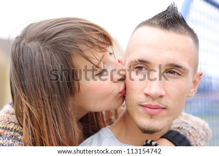 Young trendy urban couple headshot. Pretty girl with brown hair kissing boyfriend on cheek. Man has Mohawk hairstyle and earrings.