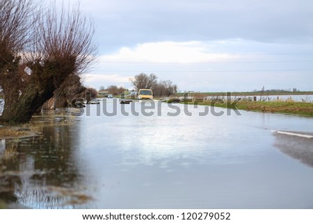 Orange van driving through flood on a rural road with flooded fields either side and cars in the background. Man walking with bicycle along bank.