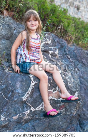 cute little girl sitting on a bid rock, wearing, colorful top, jeans shorts and pink flip flops