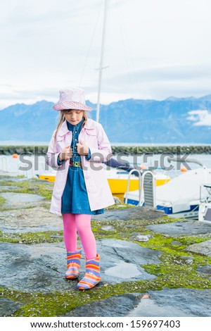 Outdoor portrait of a cute little girl, wearing pink vinyl rain coat and hat, colorful rain boots and pink tights
