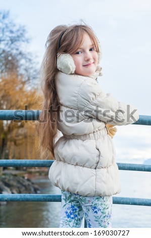 Outdoor portrait of a cute girl on a bridge on a nice winter day, wearing white jacket, printed jeans and earmuffs