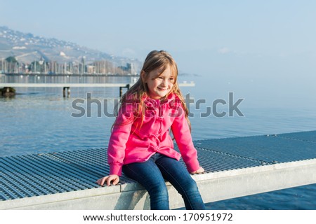 Outdoor portrait of a beautiful little girl on a nice sunny day next to lake, wearing, pink jacket, jeans and boots