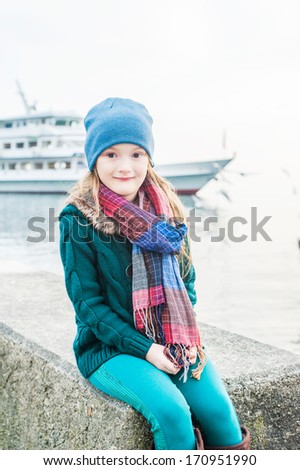Portrait of a beautiful little girl on a cold weather next to lake, wearing, green jacket and pants, hat and scarf