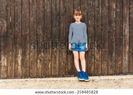 Outdoor portrait of a cute little girl, wearing frock, denim skirt and beautiful blue sneakers, standing against brown wooden wall