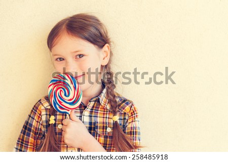 Portrait of a cute little girl of 7 years old, holding big colorful candy, wearing plaid shirt, toned image