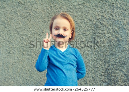 Young cute caucasian boy of 4 years old with a fake Italian moustache making a peace sign with his right hand standing in front of an old grey stone wall