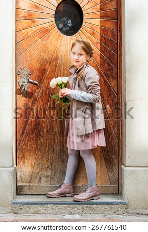Outdoor portrait of a cute little girl of 7 years old, wearing beige coat and boots, holding white roses, standing next to old wooden door