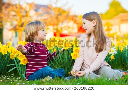 Two cute kids, little boy and his big sister, playing in the park between yellow daffodils flowers at sunset