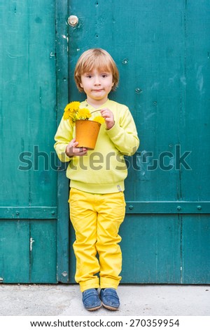 Outdoor portrait of a cute little boy, wearing yellow pullover and trousers and blue moccasins, standing by the green wooden door, holding bucket with flowers dandelions