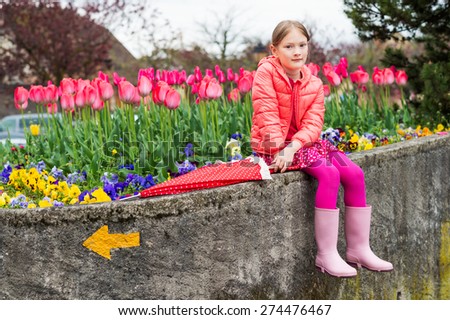 Outdoor portrait of a cute little girl, sitting next to flowerbed full of tulips, wearing bright pink clothes and rain boots, holding umbrella