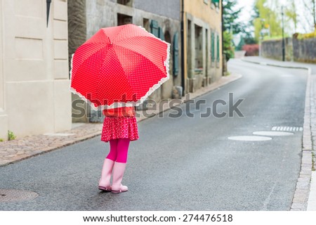 Cute little girl walking down the street, wearing pink rain boots, holding polka dot red umbrella, back view