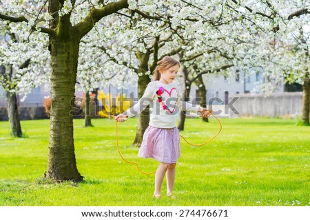 Pretty little girl playing in a spring garden on a nice sunny day, jumping with skipping rope, wearing sweatshirt with a heart and pink skirt