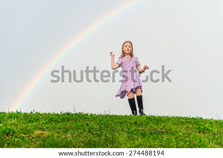 Cute little girl playing on the hill under the rainbow, wearing summer dress and black rain boots