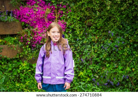 Outdoor portrait of a cute little girl under the rain, pulling her tongue, wearing purple rain coat