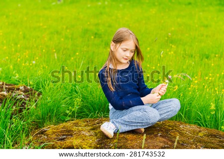 Outdoor portrait of a cute little girl resting in the nature, playing with flowers, wearing blue pullover and grey trousers