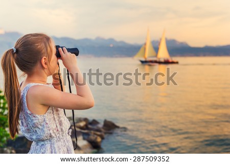 Outdoor portrait of a cute little girl playing by the lake on a nice warm evening, looking at the boat with binoculars