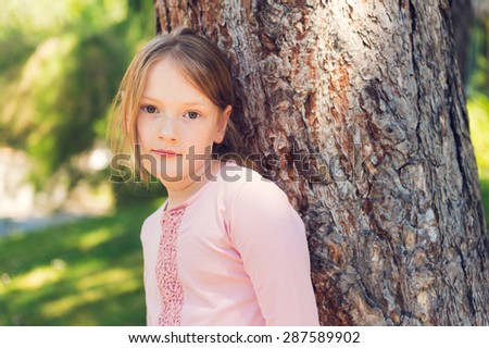 Outdoor portrait of a cute little girl of 7 years old, resting in a park, leaning on a tree