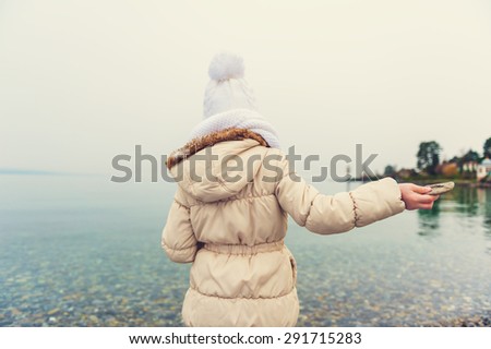 Little girl playing by the lake, throwing stones into the water on a nice cold day, wearing warm white hat, scarf and beige coat, back view, toned image