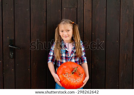 Outdoor portrait of a cute little girl holding big pumpkin with french sign Bienvenue (welcome)
