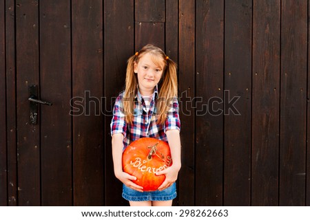 Outdoor portrait of a cute little girl holding big pumpkin with french sign Bienvenue (welcome)
