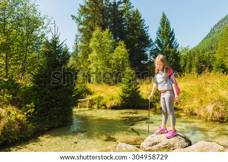 Cute little girl of 7-8 years old hiking in swiss Alps, resting by the river, wearing sport clothes, trainers and backpack