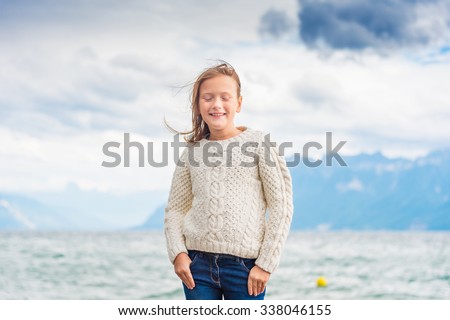 Cute little girl of 8 years old playing by the lake on a very windy day, wearing warm white knitted pullover