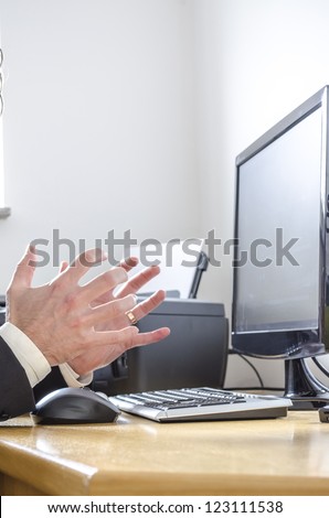 Businessman at office desk making angry gesture with hands towards computer.