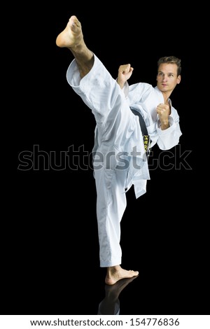 Young man in white kimono training karate kicking his leg at you. Isolated over black background.