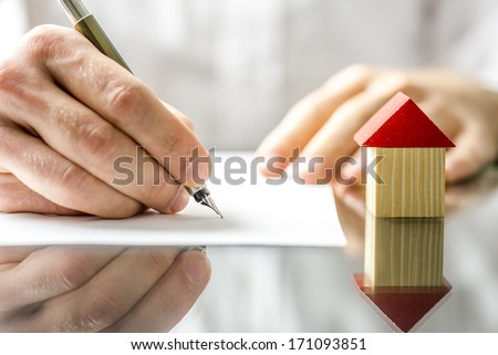 Conceptual image of a man signing a mortgage or insurance contract or the deed of sale when buying a new house or selling his existing one with a small wooden model of a house alongside