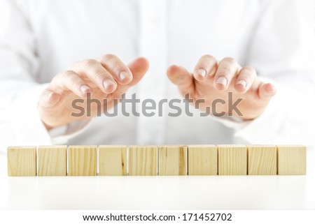 Man holding protective hands above a line of ten blank wooden cubes as he safeguards and protects them. Ready for your text.