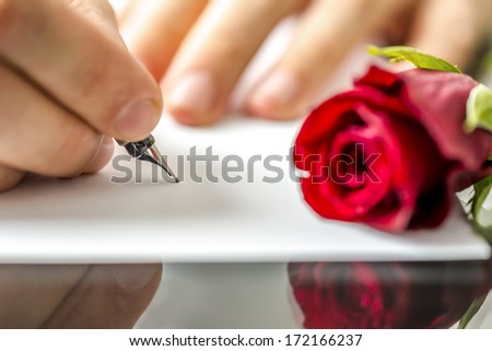 Man writing a letter to his sweetheart or lover for Valentines or an anniversary with a red rose lying alongside the notepaper, view of his hands
