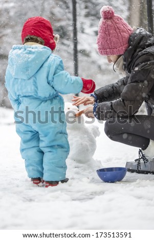 View from the rear of a young mother and toddler working in the fresh white snow in the garden building a winter snowman
