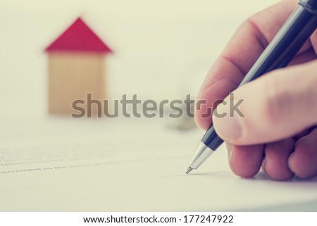 Retro image of a man signing a deed of sale, mortgage document or insurance contract on a house with a closeup view of his hand with a small wooden model of a house.