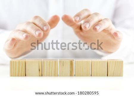 Man holding protective hands above a line of seven blank wooden cubes as he safeguards and protects them. Ready for your text.